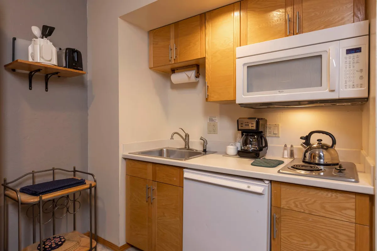 The image presents a warmly lit kitchen interior featuring modern appliances, wooden cabinetry, a spacious countertop, and a sink. The layout invites both cooking and casual interactions, with subtle cues of morning or evening activity such as a coffee maker and utensils in use.