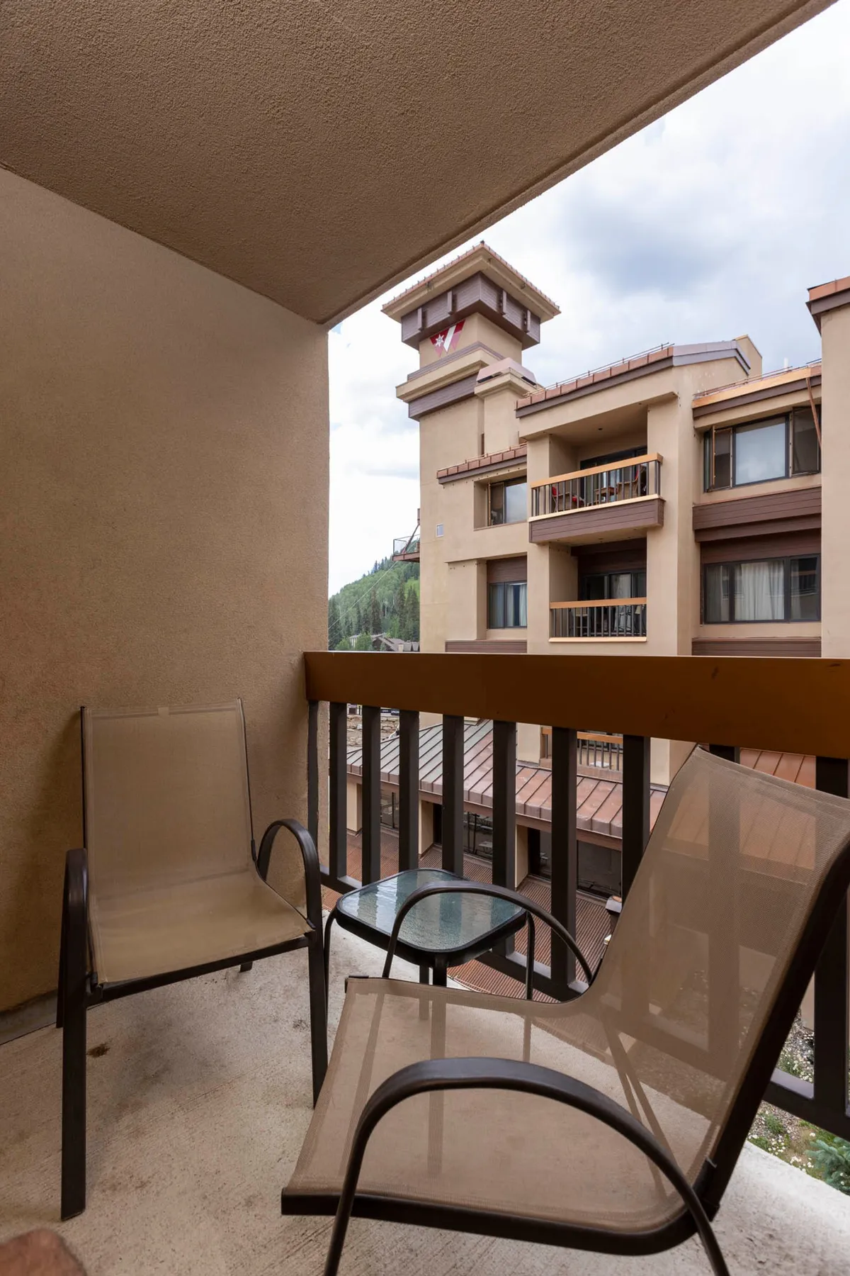 This image captures a serene balcony setting with chairs and a glass table overlooking surrounding alpine buildings and stunning mountain views. The soft natural light and tranquil atmosphere create the perfect spot for morning coffee or intimate evening conversations while taking in the breathtaking landscape.