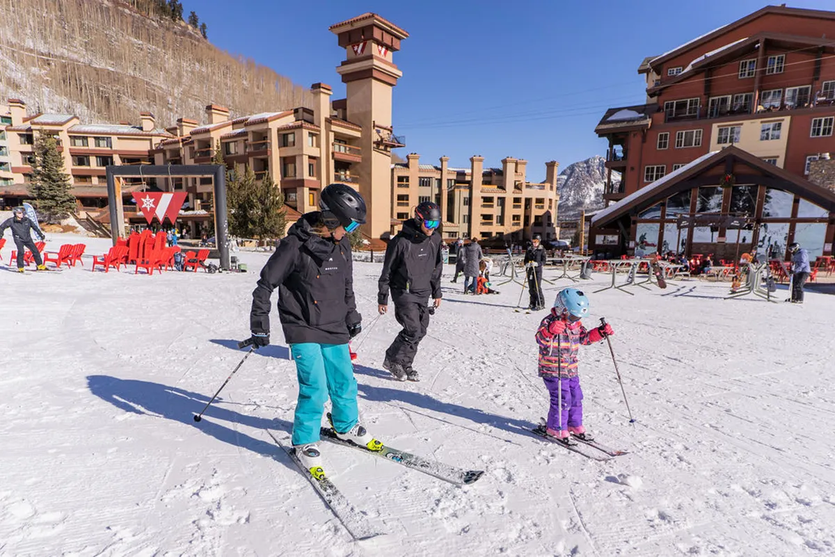 A family skis together near resort buildings, surrounded by a snow-covered landscape. The scene exudes playful energy and highlights winter sports as a memorable family bonding experience.