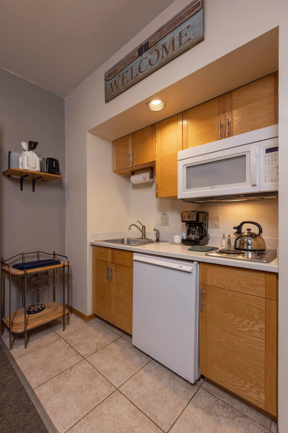 This cozy kitchen scene highlights a 'WELCOME' sign, contemporary appliances, wooden cabinets, and a thoughtfully arranged countertop. The mood conveys warmth and sociability, with hints of people cooking and sharing food and drinks.