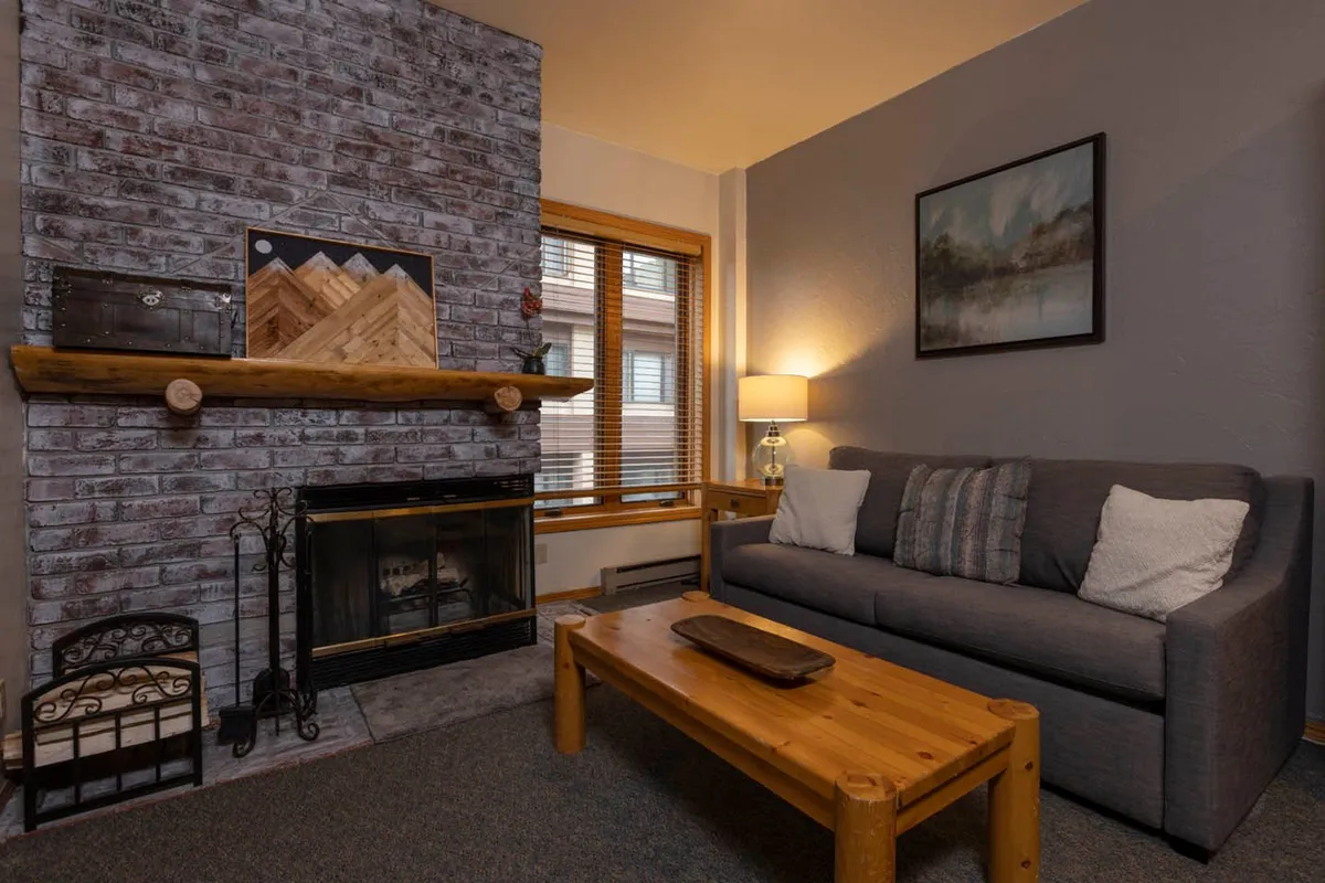 The image showcases a cozy living space featuring a classic brick fireplace as the centerpiece, a wooden coffee table set in front, a plush sofa adorned with cushions, and tasteful mountain-themed artwork decorating the wall. The scene radiates warmth, inviting guests to relax by the fire, enjoy a cup of hot cocoa, or share intimate conversations in a welcoming mountain retreat.