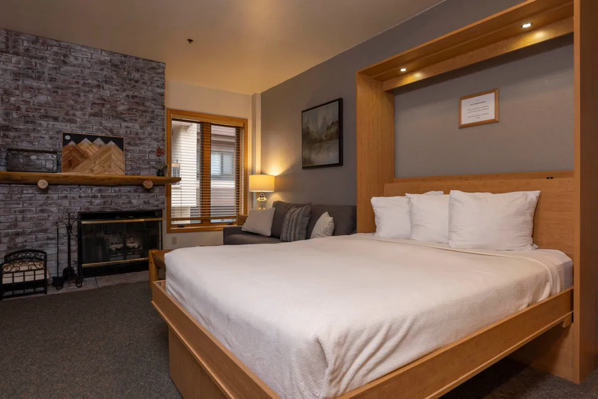 This image presents a cozy bedroom interior featuring a luxurious bed with plush bedding, a glowing fireplace, a soft sofa, and tasteful artwork on the walls. The scene radiates comfort and tranquility, encouraging relaxation and restful moments.