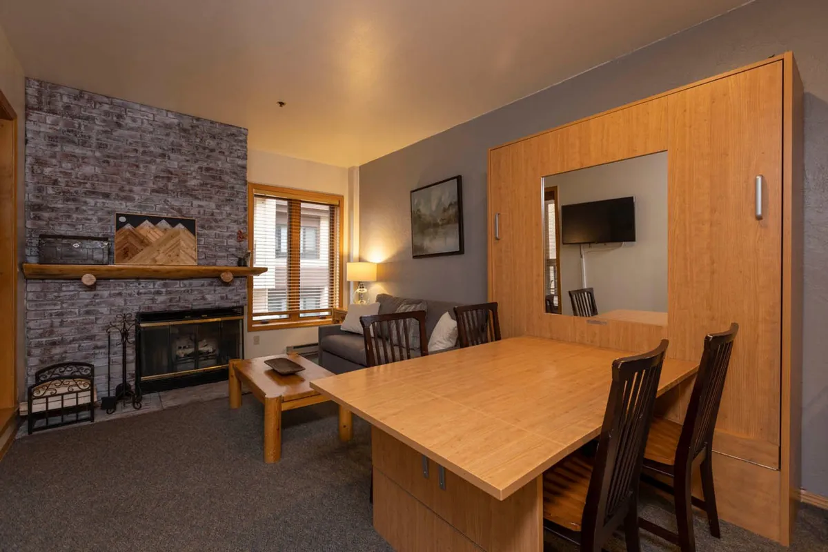This image displays a warm, inviting living room featuring a prominent wood-burning fireplace, a comfortable sofa, a dining table, and tasteful artwork on the wall. The space is styled to encourage relaxation and togetherness, with the fireplace as the anchoring focal point between the living and dining areas.