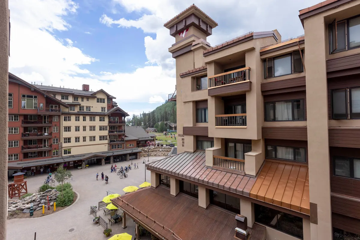 A sunlit, bustling hotel courtyard with people relaxing under yellow umbrellas, outdoor seating, and bikes nearby. The atmosphere is lively, with guests socializing and soaking up the mountain village vibe.
