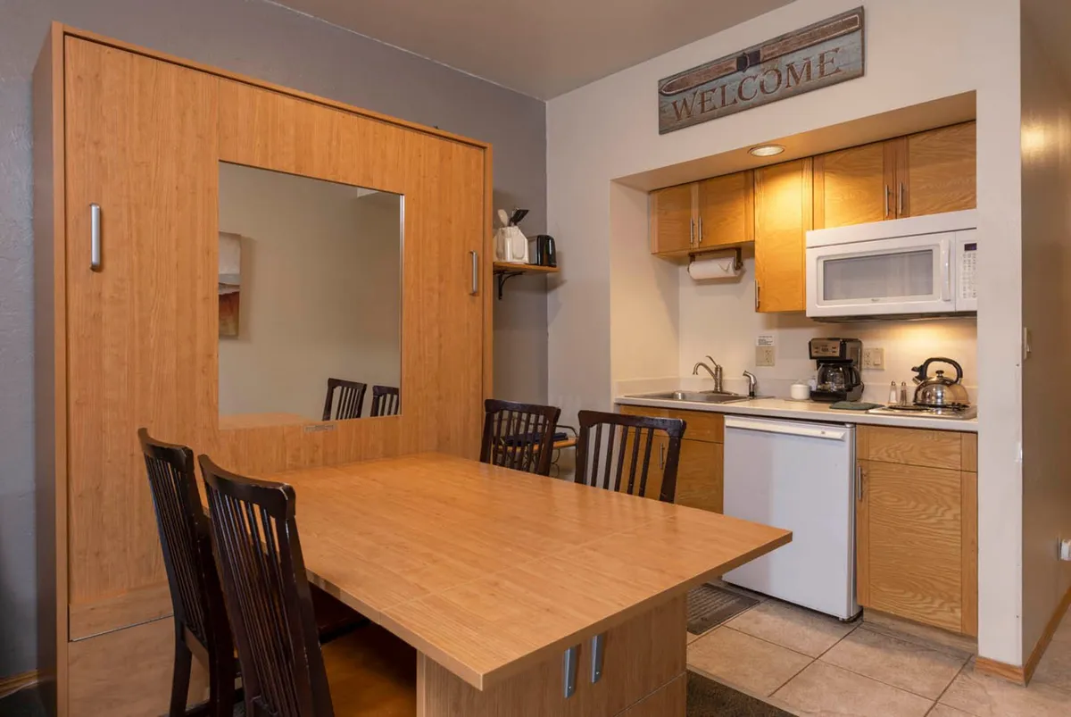 The scene centers on a welcoming dining table set within an open interior, with views to a nearby kitchen area and a mirror cabinet. A prominent 'welcome' sign and warm lighting enhance the sense of hospitality and togetherness, reinforcing the communal gathering aspect.