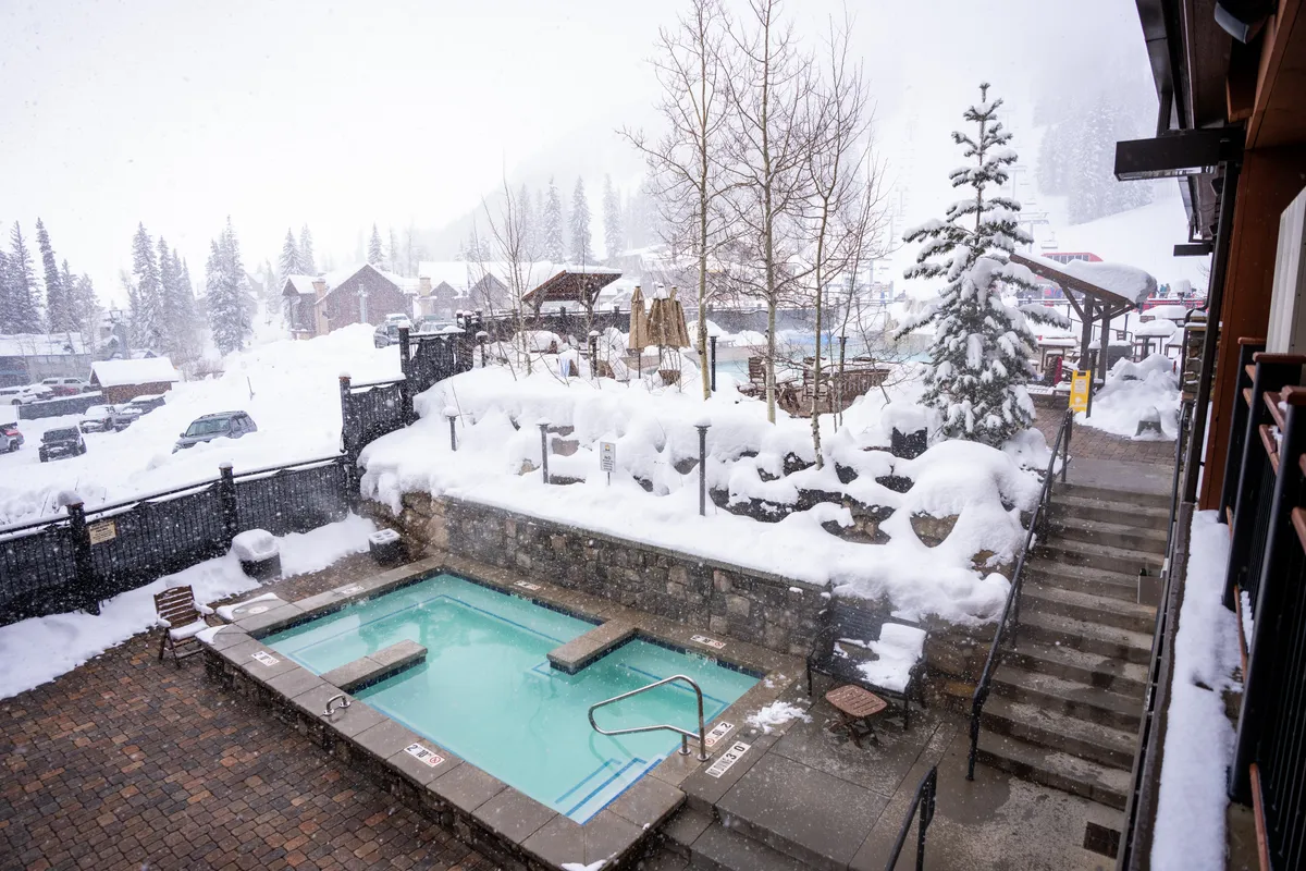 This image captures a rooftop hot tub surrounded by snow-covered landscape and trees. The scene is tranquil and inviting, with the warm steam rising from the water contrasting beautifully with the winter scenery. Steps lead up to the hot tub, emphasizing accessibility, and the overall mood highlights relaxation and luxury in a mountain setting.