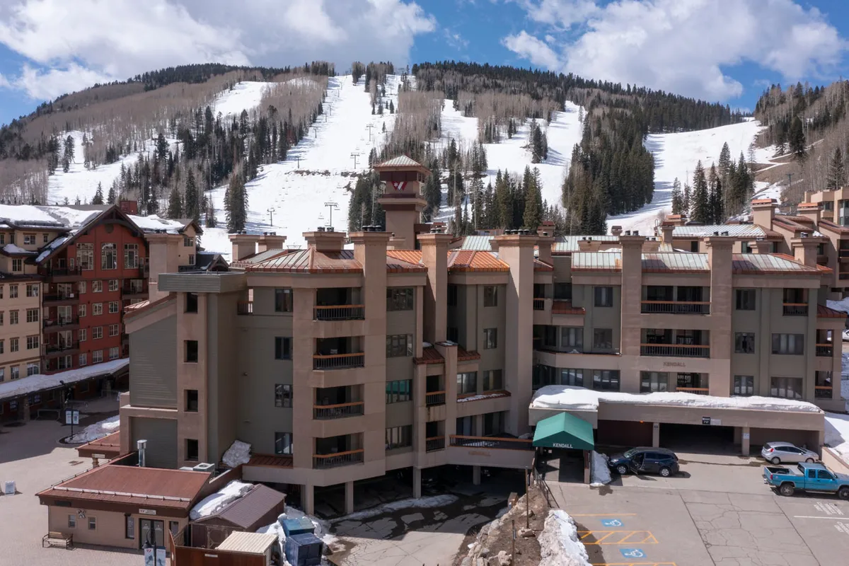The building facade of the resort stands against breathtaking snowy mountain slopes, with a visible parking area and hints of outdoor activity. The vista blends the spirit of adventure with the comfort and accessibility of the accommodations.