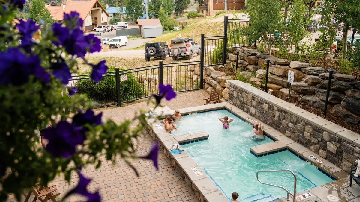 An inviting outdoor heated swimming pool is surrounded by flowers and stone walls, with children playing in the water and people relaxing poolside. The image conveys a joyful, social atmosphere and the pool’s integration into the natural landscape, making it ideal for both recreation and relaxation.